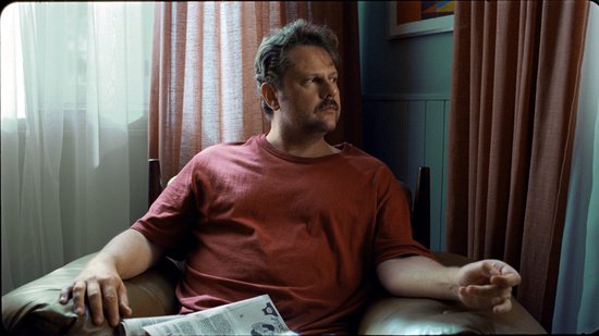 Man met snor in een rode T-shirt, zittend in een bruine fauteuil met een krant.