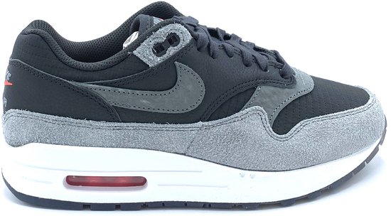 Zij-aanzicht van zwarte Nike Air Max 1 sneaker, met grijze suède en lederen overlays, witte zool en rode Air unit.