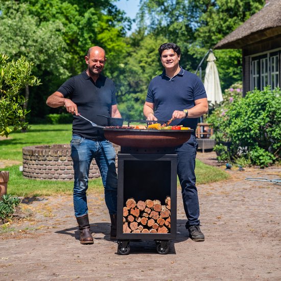 Twee mannen bij een grote zwarte barbecue met houtopslag. Een man met schort en jeans, de ander in een blauw poloshirt.