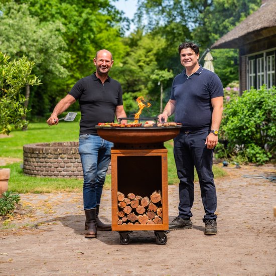 Twee mannen staan naast een grote ronde barbecue op wielen, gevuld met gegrild eten en houtblokken in de opslag onderaan.