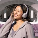 Een vrouw met gesloten ogen, genietend van muziek met een lichtgrijze draadloze on-ear koptelefoon, met festivalpodium op achtergrond.