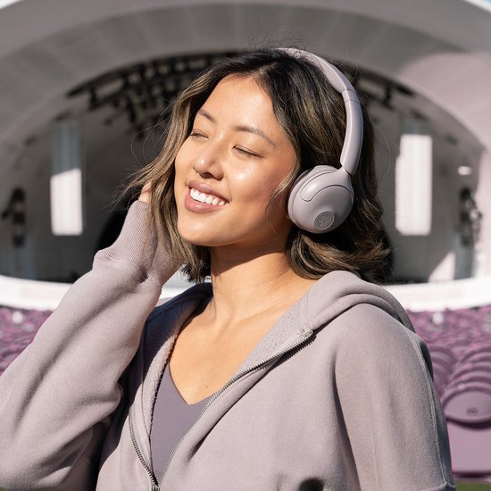 Een vrouw met gesloten ogen, genietend van muziek met een lichtgrijze draadloze on-ear koptelefoon, met festivalpodium op achtergrond.