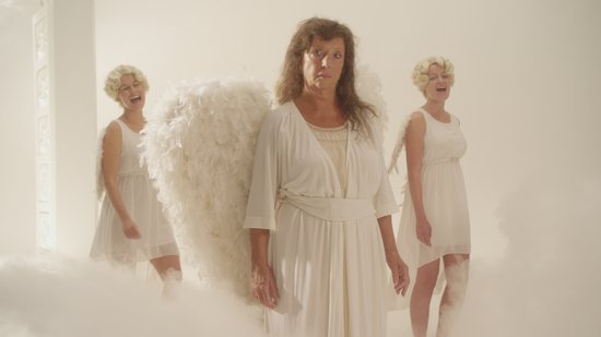 Drie vrouwen in witte jurken, twee met blonde krulbojes en vleugels, de middelste vrouw met grote witte engelen vleugels en bruin haar.