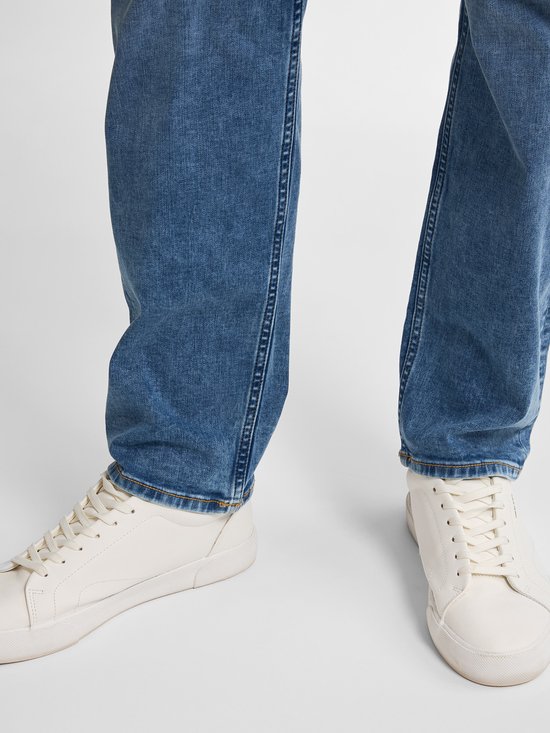 Close-up van de onderbenen van een man in een lichtblauwe jeans en witte sneakers.