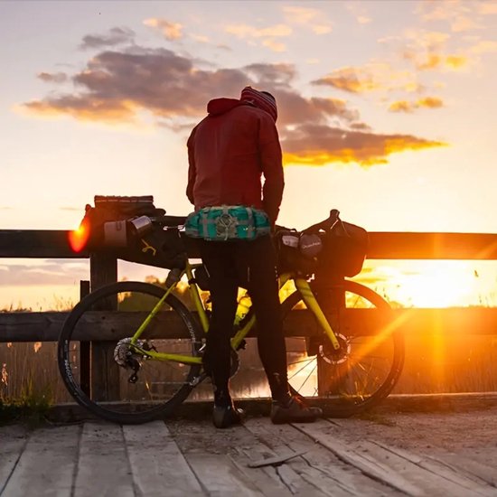 Man met rode jas en groene heuptas staat bij een fiets bij zonsondergang.