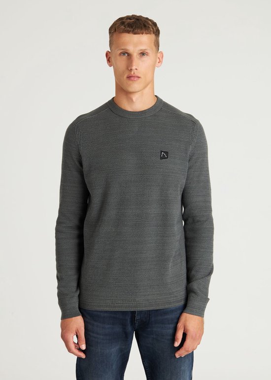 Homme portant un pull tricoté gris foncé avec un petit écusson noir et logo blanc sur la poitrine gauche.