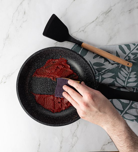 Een hand schuurt tomatensaus uit een koekenpan met marmerlook antiaanbaklaag. Een zwarte spatel ligt ernaast op een groen blad.
