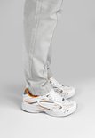 Voet van model met witte DUTCH'D sneakers met oranje en grijze accenten, onder een grijze broek.