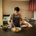 Man in schort kookt pasta met spekjes in een zwarte koekenpan met granieten look.