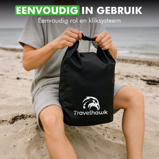 Een persoon op het strand, zittend, houdt een zwarte Travelhawk drybag vast. De tas heeft een rolsluiting.
