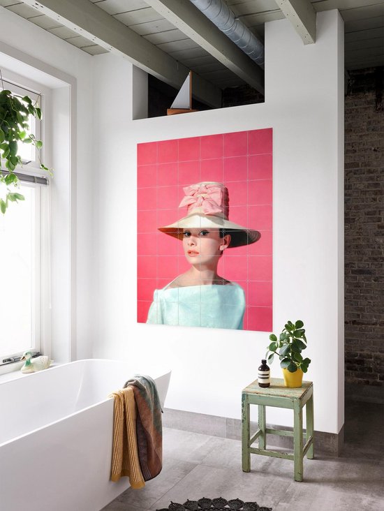 Audrey Hepburn portant un grand chapeau, un nœud rose et un haut bleu pâle, sur fond rose, dans une salle de bain près d'une baignoire.