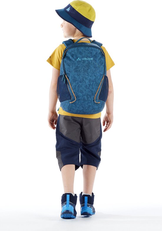 Jongen met gele hoed en rugzak, staand met de rug naar de camera. De rugzak is blauw met een geometrisch patroon.