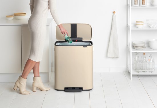 Une femme en robe en tricot jette une bouteille dans la poubelle Brabantia Bo crème dans une cuisine.