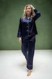 Model draagt een donkerblauwe satijnen pyjama met witte biesjes, staand op blote voeten op een witte vloer.