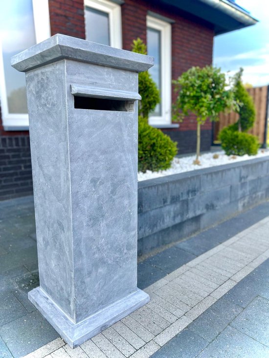 Grijze hardstenen brievenbus met een gleuf aan de voorzijde, naast een woning met een groen aangelegde border.