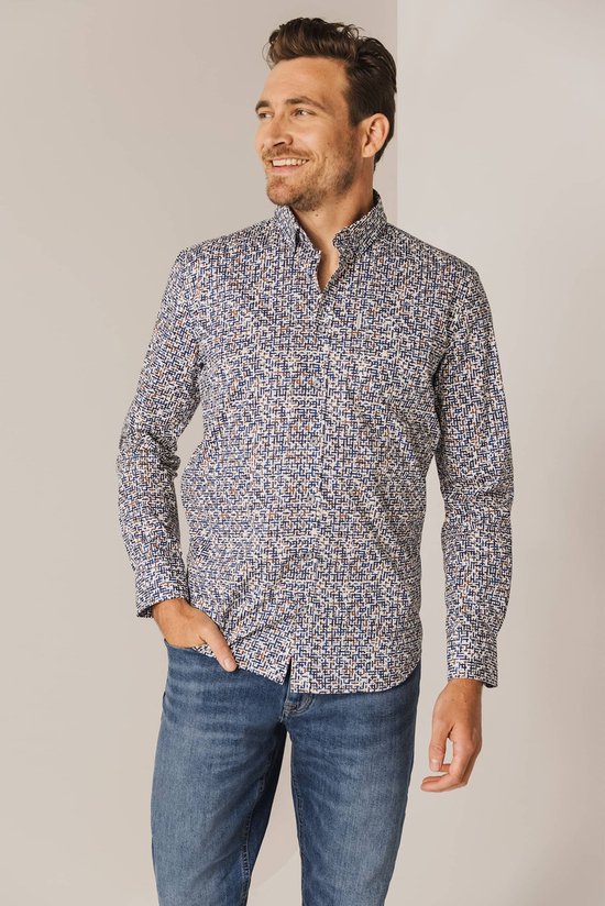 Man met hand in zijn broekzak, draagt een blauw overhemd met een witte, bruine en donkerblauwe print. Hij draagt een jeans.