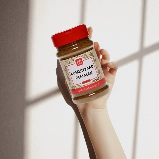 Hand houdt pot gemalen komijnzaad met rode dop vast. Het etiket toont de tekst 'KOMIJNZAAD GEMALEN'.