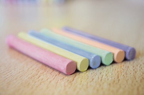 Een rij van zeven gekleurde krijtjes: roze, geel, lichtblauw, groen, oranje, beige en paars, liggend op een houten ondergrond.