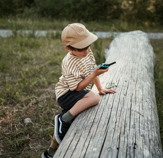 Jongetje met pet, zittend op een boomstam, kijkend naar een blauwe Kidywolf KIDYTALK walkietalkie.