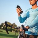 Een man met een blauwe fietsoutfit houdt een zwarte sportfles vast naast zijn racefiets.