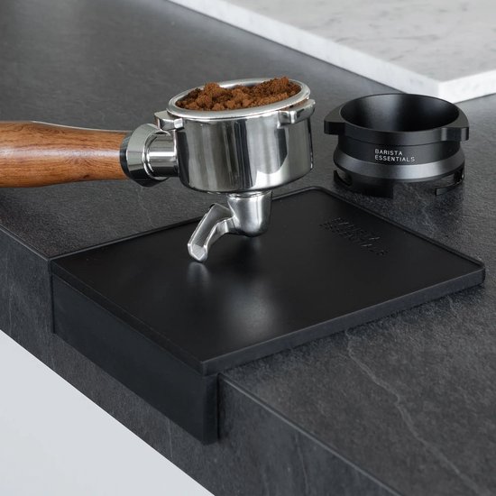 Een portafilter met koffie en een doseerring, staand op een zwarte tampermat op een donker werkblad.