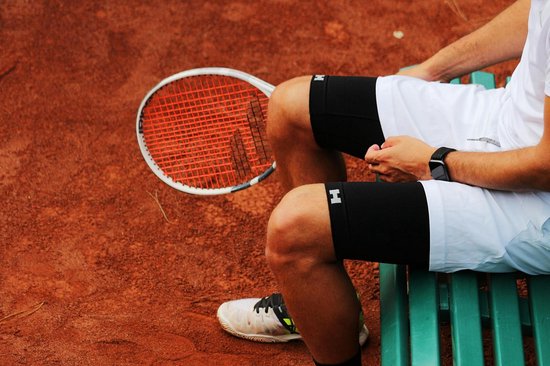 Man op een tennisbaan, zittend met een witte korte sportbroek en zwarte compressieshorts.
