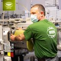 Man met mondkapje in groene T-shirt met logo van Bekina Boots, werkt aan machine in fabriek. "Made in Belgium Since 1962" logo.