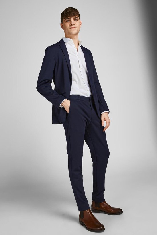 Man met donkerblauwe blazer en broek, wit overhemd en bruine schoenen.