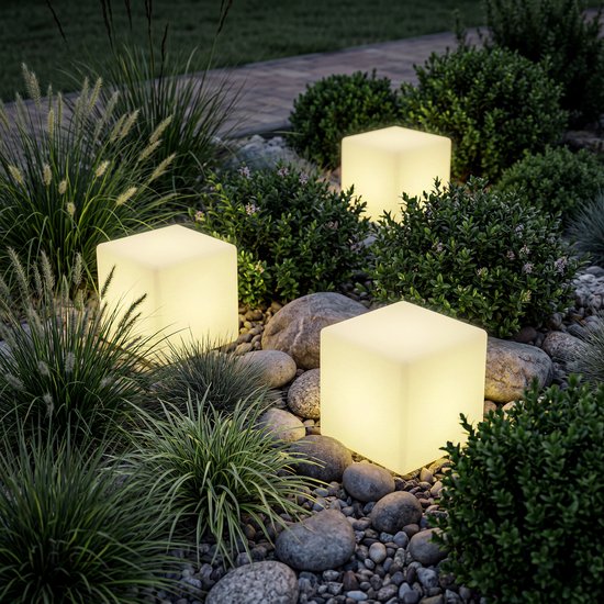 Drie verlichte witte kubuslampen in een tuin tussen planten en stenen.