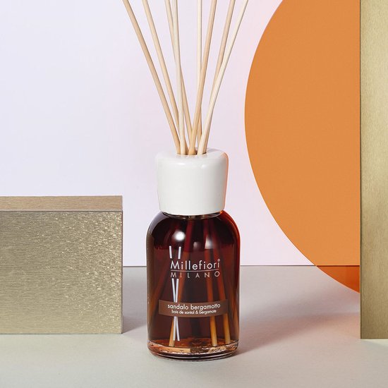 Bruine glazen fles diffuser met witte dop, stokjes en een gouden kubus en oranje cirkel op de achtergrond.