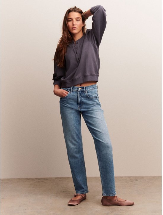 Model met een grijze sweater en blauwe mom jeans met rechte pijpen, staand met de hand in de zak.