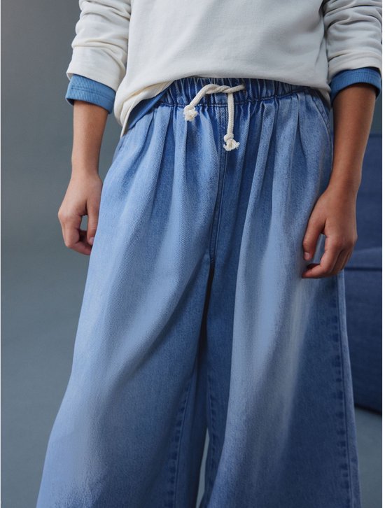 Kind draagt wijde, ronde jeans in lichtblauw denim, met strik aan de tailleband.