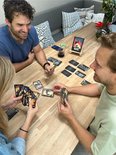 Drie personen spelen een kaartspel aan een houten tafel. Een tablet toont het logo "SLA'A".