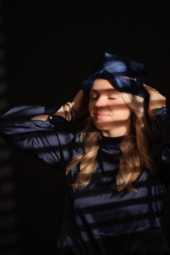 Een vrouw met blond haar met gesloten ogen draagt een donkerblauw zijden slaapmasker van Slowwave.