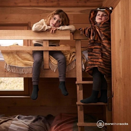 Twee kinderen in een houten huisje, één zit op een bed en de ander staat op een trapje, beiden dragen zwarte sokken.