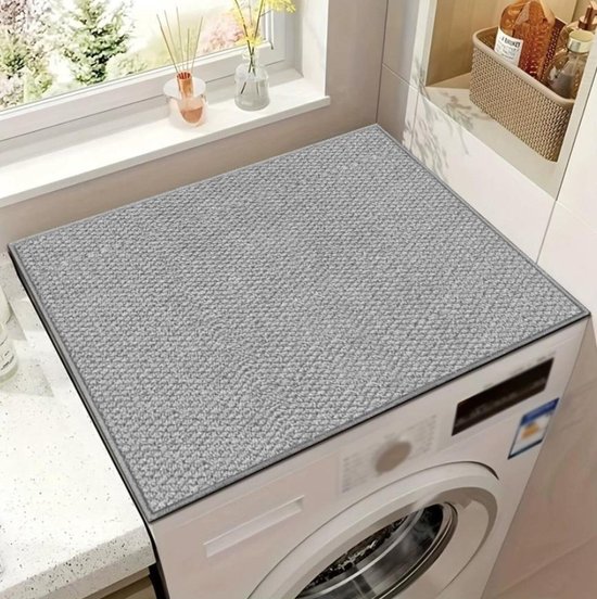Grijze wasmachinemat met een geweven textuur, geplaatst op een wasmachine naast een raam.