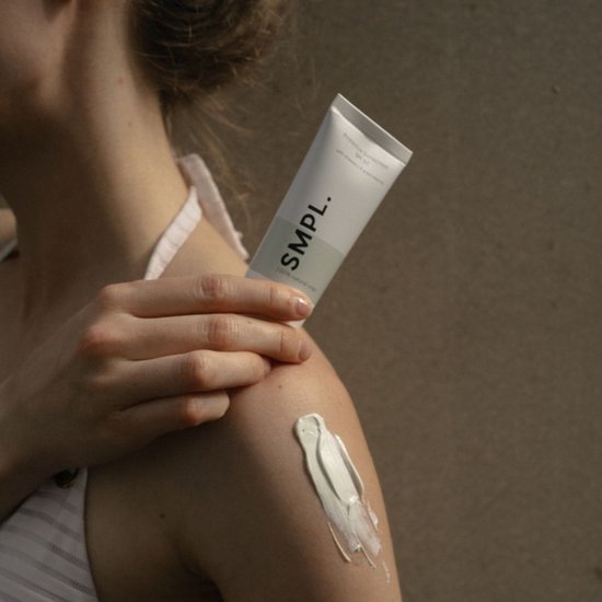 Een vrouw brengt witte zonnebrandcrème aan op haar schouder. De tube heeft de tekst "SMPL. Protective Sunscreen SPF 30".
