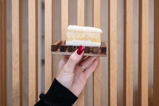 Een stuk loofah zeep met een laagje honing en melk op een houten zeepbakje, vastgehouden door een hand met rode nagellak.