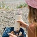 Crème VANN drinkfles met zilveren onderkant, vastgehouden door een vrouw op het strand.