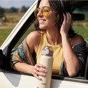 Crème VANN drinkfles met zilveren onderkant, vastgehouden door een vrouw met zonnebril in een auto.