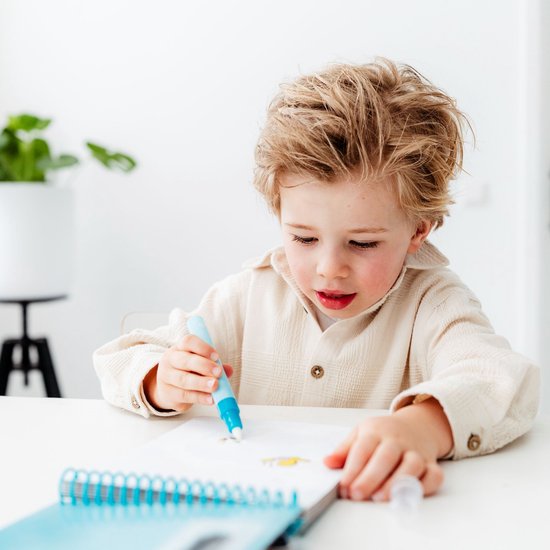 Jongen met blond haar die in een notitieboekje tekent met een blauw-witte pen, hij draagt een crème kleurig shirt.