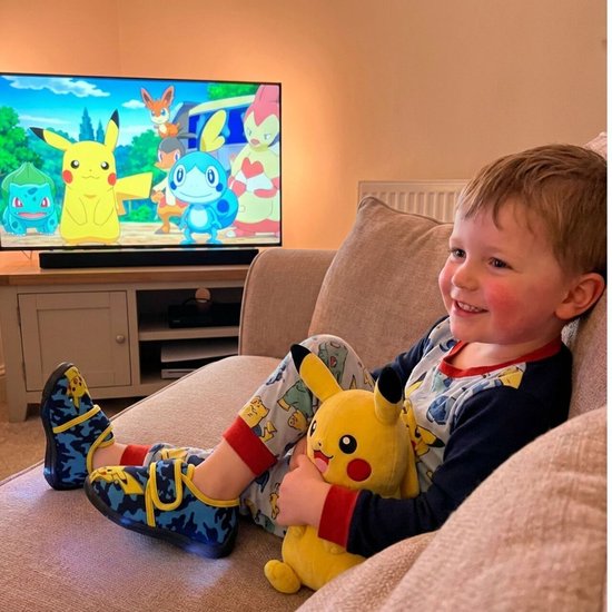 Jongen zit op een bank met een Pikachu knuffel, kijkend naar tv. Hij draagt pantoffels met Pikachu erop.