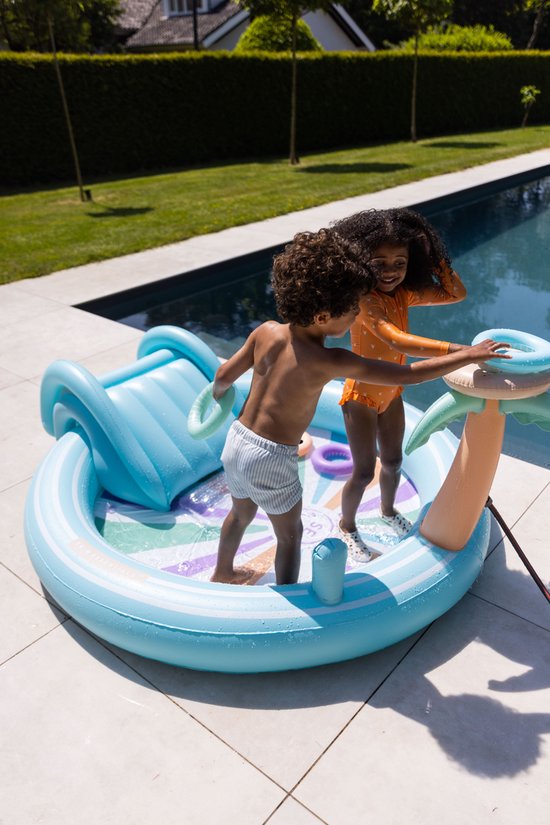 Kinderen spelen in een opblaasbaar zwembad met glijbaan en palmtak sproeier. De bodem heeft een zonnespatroon met gekleurde strepen.
