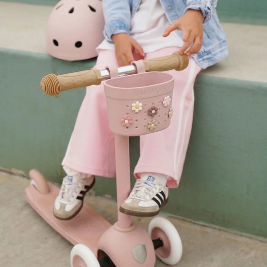 Een roze Banwood kinderstep met een mandje met bloemen erop, een meisje zit erop met een helm.