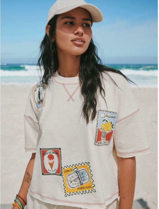 Vrouw met donker haar in een crème T-shirt met vakantie-emblemen voor een blauwe oceaan.