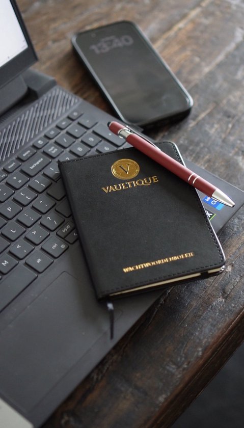 Zwart wachtwoordenboekje met gouden 'V' logo van Vaultique, een rode pen en een smartphone op een laptop.