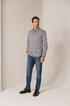 Man met een blauw-wit met bruine print overhemd en spijkerbroek, staand met hand in zak.