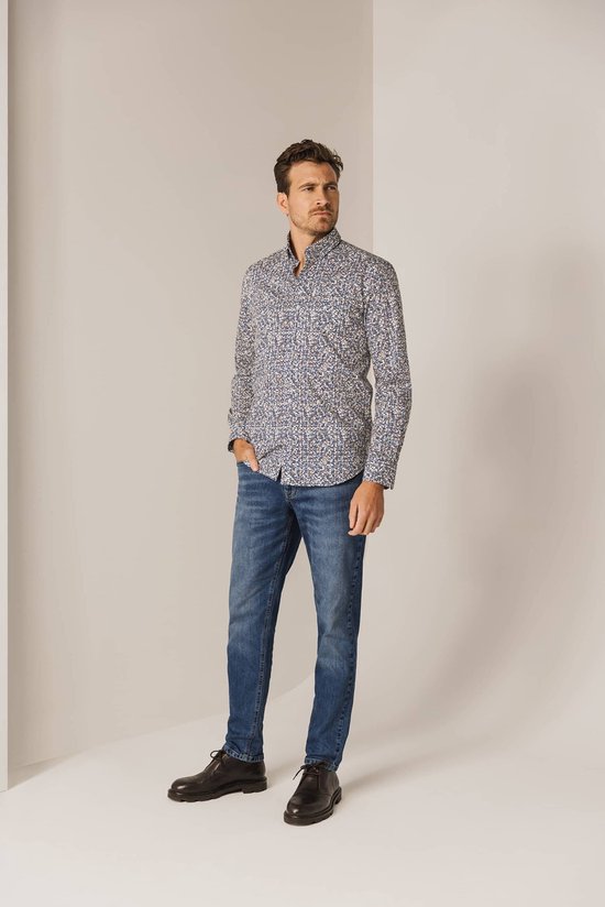 Man met een blauw-wit met bruine print overhemd en spijkerbroek, staand met hand in zak.