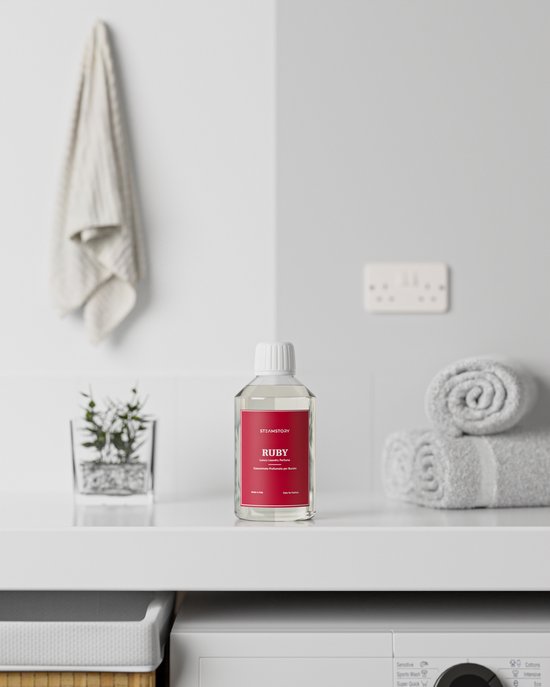 Fles SteamStory "Ruby" wasparfum op een aanrecht in een badkamer, met handdoeken en plantje.