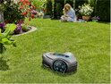 Grijze robotmaaier met turkooizen accenten op een groen gazon, vrouw leest boek op de achtergrond.
